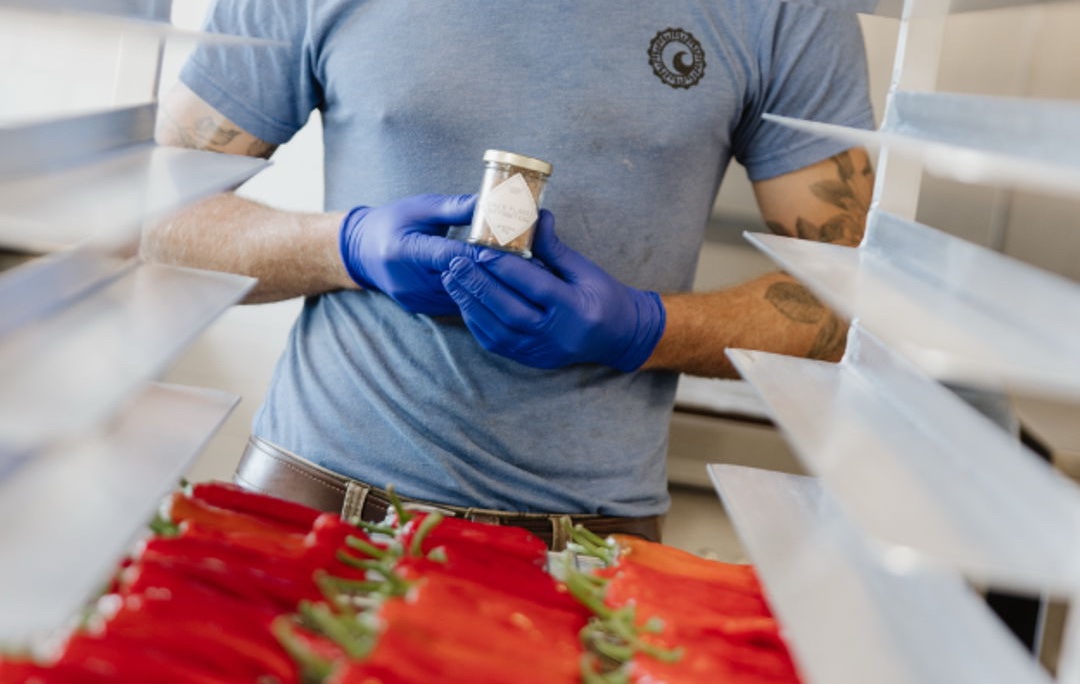A Maker in blue gloves holding a jar beside racks of red peppers